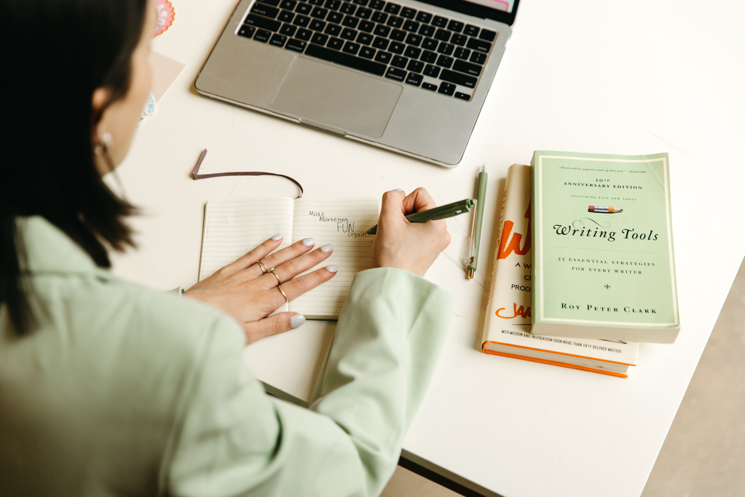 Over-the-shoulder view of a woman in a mint-green blazer writing “Make Marketing FUN again” in a notebook next to a laptop and a stack of books, including Writing Tools by Roy Peter Clark. The scene captures a moment of strategic brainstorming aligned with the blog’s message about ditching corporate jargon in favor of clear, compelling storytelling for disruptive brands.